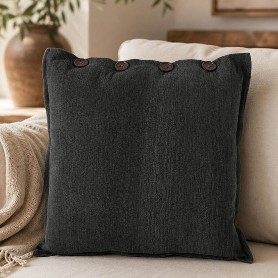 Sofakissen schwarz „Joscha“ von Boltze auf einem cremefarbenen Sofa, dunkle Knöpfe an der Oberkante, daneben eine Strickdecke und eine Tonvase