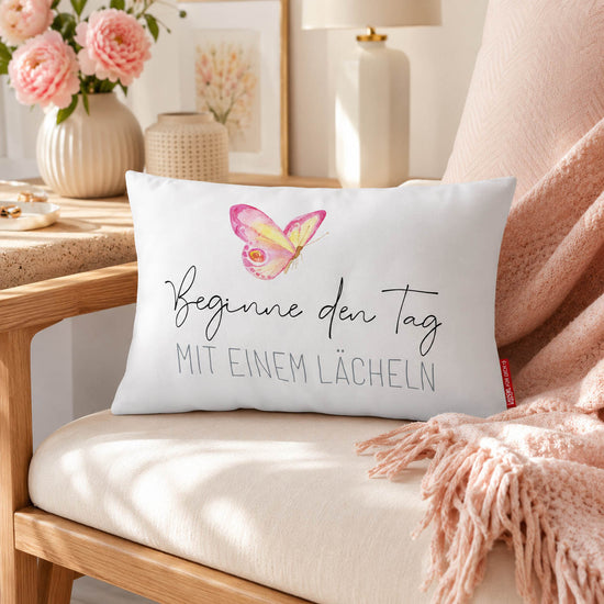 Kissen mit Spruch Beginne den Tag mit einem Lächeln mit Schmetterling und rosa Rückseite auf hellem Bettzeug